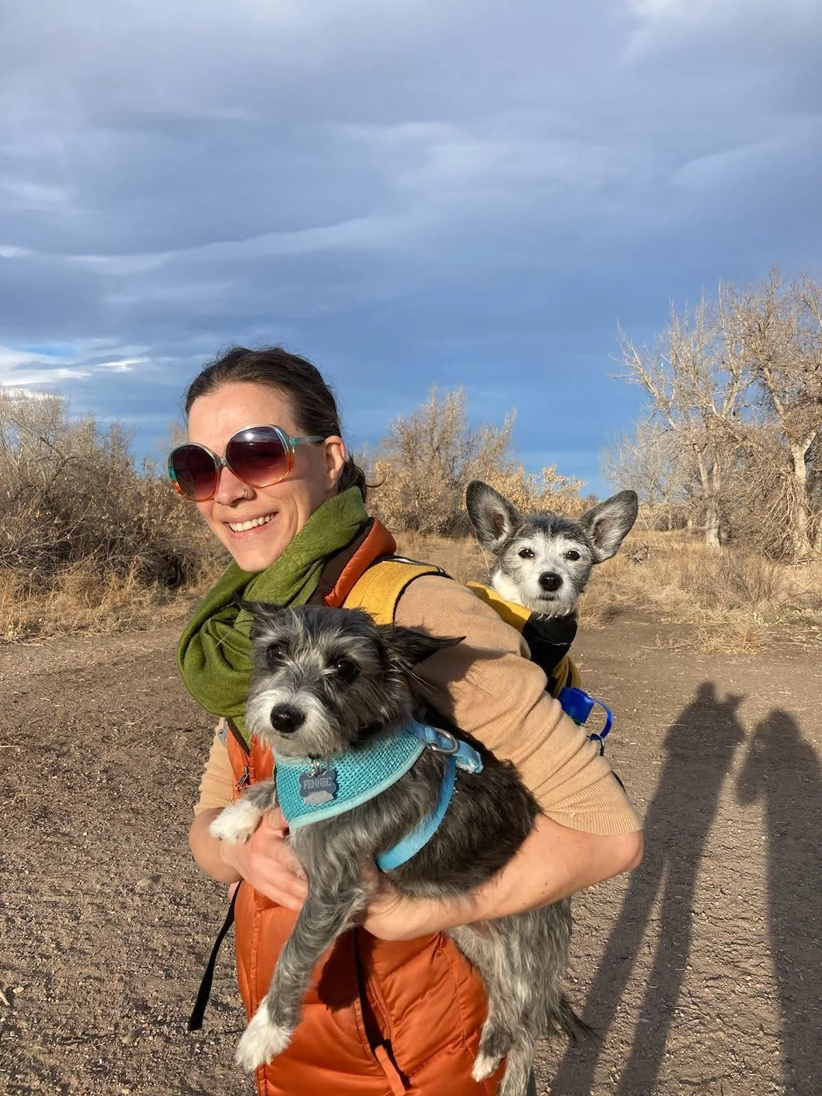 A woman wearing sunglasses and colorful clothes holding two small dogs outdoors on a dirt trail in a dry, leafless forested area under a cloudy sky.
