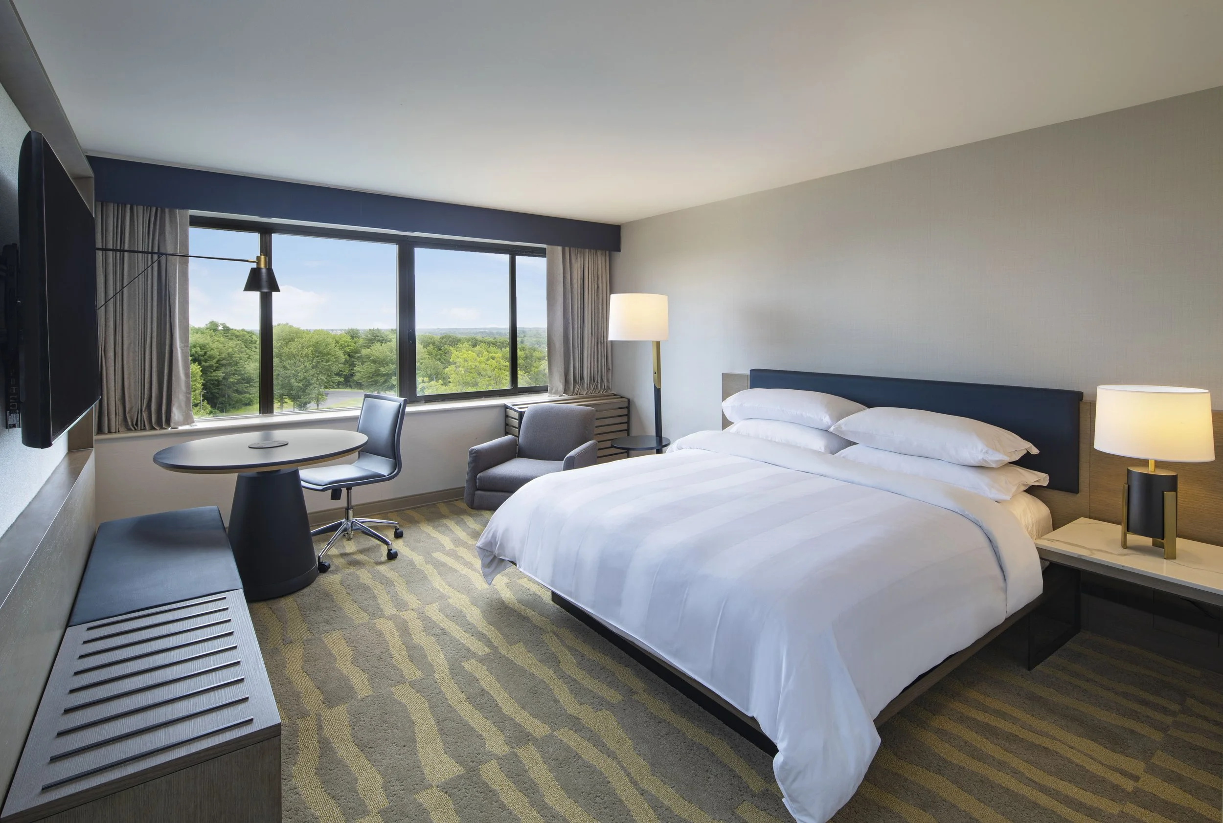 Modern hotel lobby with a spiral staircase, large windows with sheer curtains, various black and gray sofas, armchairs, and a round coffee table with decorations. Art piece on the wall and a vase on a side table.