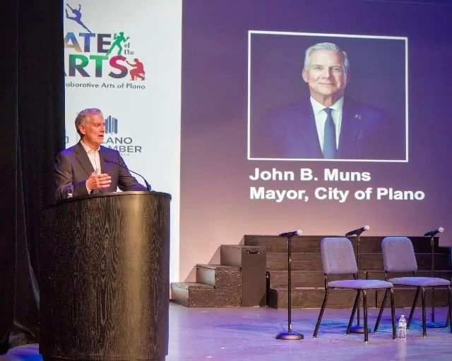 A man in a suit standing at a podium on stage giving a speech. Behind him is a large screen displaying a portrait of John B. Muns, Mayor of the City of Plano, along with the logo for the Arts of the Arts.