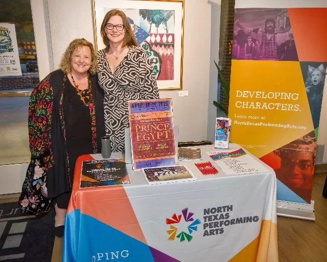 Two women standing behind a table with North Texas Performing Arts promotional materials, in an art gallery setting.