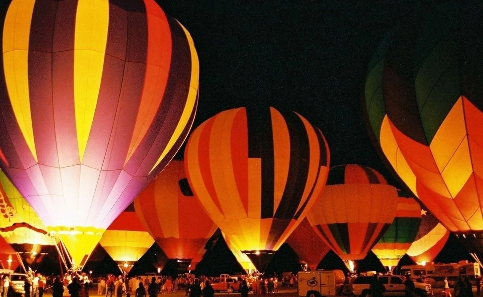 Colorful hot air balloons illuminated at night with crowds of people nearby.