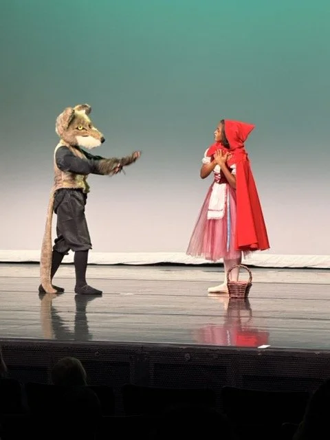 A person in a wolf costume talking to a young girl dressed as Little Red Riding Hood on stage.