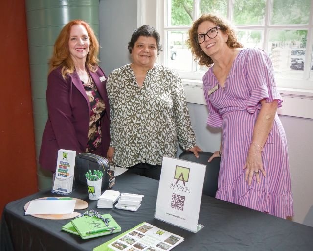 Three women standing behind a table at an indoor event, smiling for the camera. The table has promotional materials, a sign with a QR code, and some green notebooks. The background features a window with a view of trees.