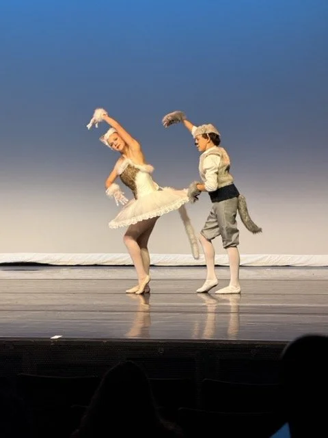 Two ballet dancers in costume perform on stage against a blue background. One dancer, wearing a white dress with a tutu, is spinning with arms raised. The other, dressed as a mouse, stands behind her holding her tutu.