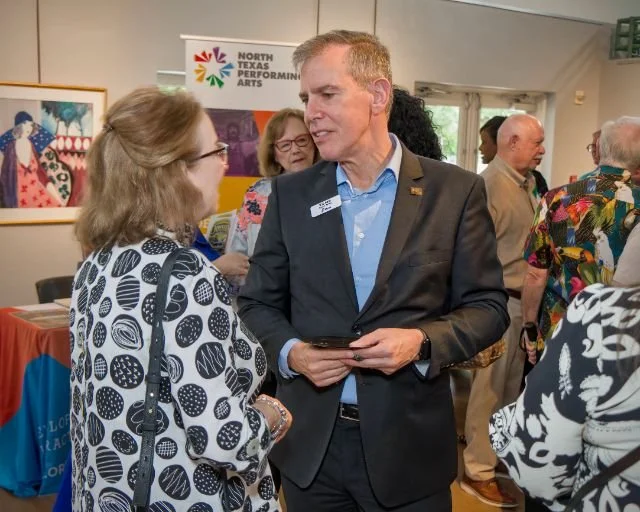 A man in a suit and a woman with glasses and patterned blouse are engaged in conversation at an indoor event. There are other people and a banner for North Texas Performing Arts in the background.