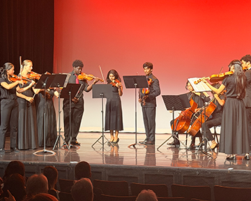 Group of young musicians performing on stage with string instruments, dressed in black and blue formal attire, under red stage lighting.