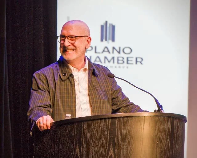 A man with glasses and a beard stands behind a podium, smiling, with a microphone attached. Behind him is a screen with a logo and the text 'Plano Chamber.'