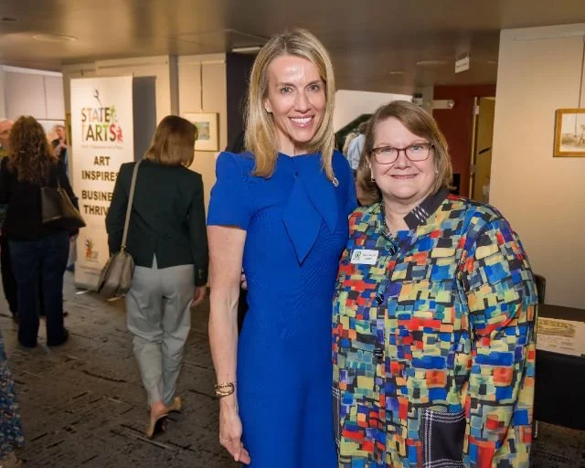 Two women standing together at an indoor event, smiling at the camera. One woman is wearing a blue dress, and the other is dressed in a multicolored patterned jacket with a name tag. In the background, other attendees are visible, along with a poster
