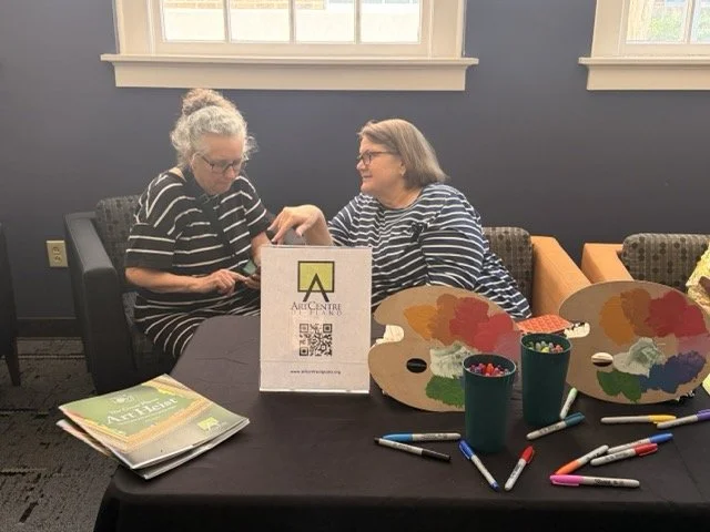 Two women sitting at a table, engaging in an art activity, with paint palettes, markers, and educational materials on the table, inside a room with dark blue walls and a window.