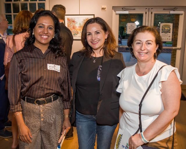 Three women standing together indoors at a social event, smiling at the camera. The woman on the left has dark hair, glasses, and is wearing a striped shirt and patterned pants. The woman in the middle has shoulder-length hair, a black jacket, and je