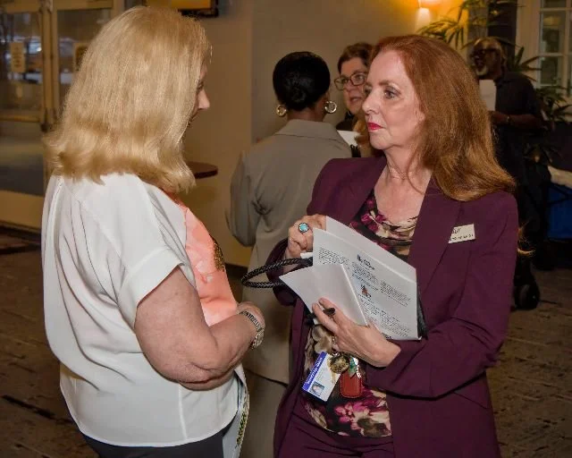 Two women engaged in conversation at an indoor event, one holding a brochure and wearing a purple blazer with a name tag, and the other with blonde hair wearing a light-colored shirt, with several people in the background.