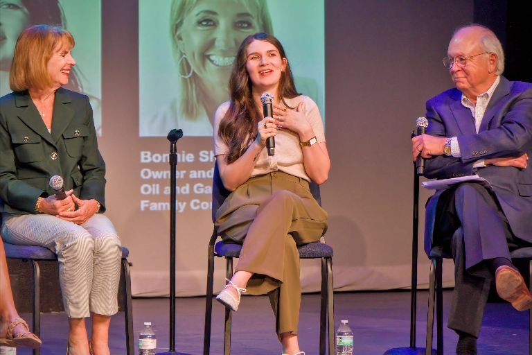 Three people sitting and talking on stage during a panel discussion, with a large screen behind them displaying a woman's face and some text.