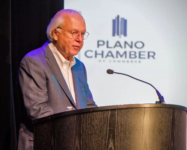 An older man with white hair and glasses stands at a podium with a microphone, in front of a screen displaying the Plano Chamber of Commerce logo.