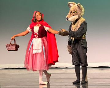 A woman dressed as Little Red Riding Hood holding hands with a person in a wolf costume on stage.