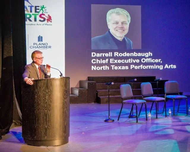 Man in a suit speaking at a podium during a presentation with a large screen behind him displaying a photo of Darrell Rodenbaugh, Chief Executive Officer of North Texas Performing Arts, and event logos.
