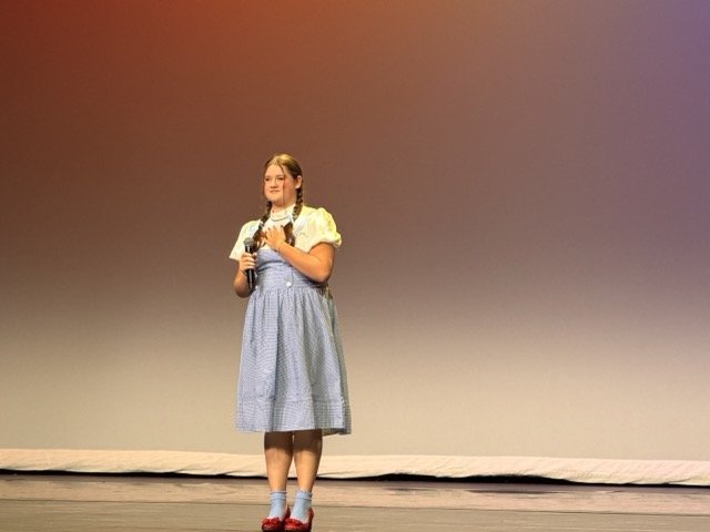 Young girl standing on stage holding a microphone, wearing a blue dress with a white blouse, and red shoes with socks, in front of a plain gradient background.