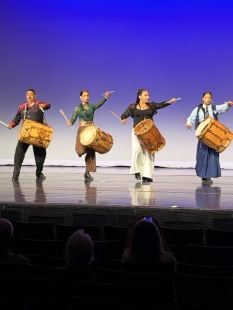 Five performers on stage playing traditional drums, dressed in colorful costumes, under blue stage lighting.