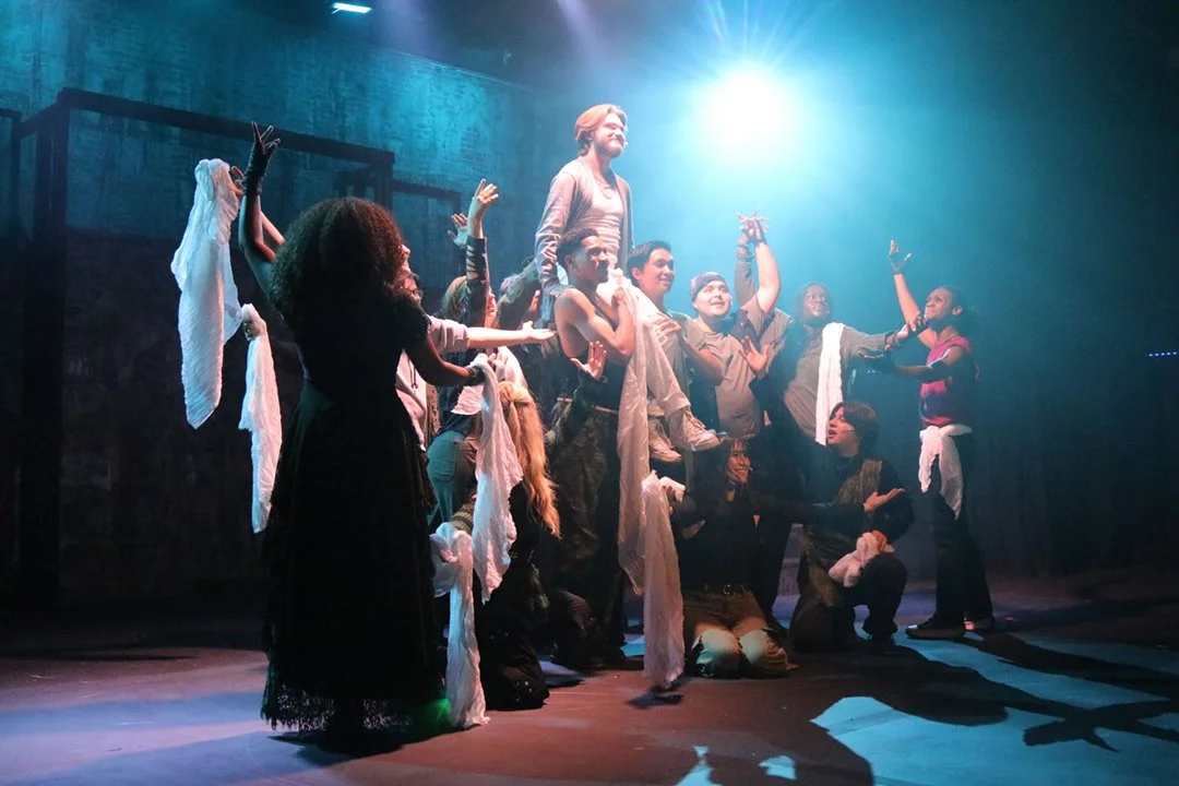 Group of performers on stage, with one person elevated, under dramatic lighting and blue background.