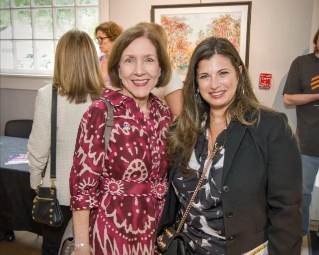 Two women smiling at an indoor gathering, with other people blurred in the background, and a painting on the wall behind them.