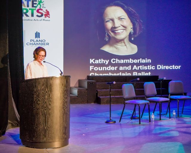 A woman standing at a podium on stage giving a speech, with a large screen behind her displaying her name, Kathy Chamberlain, and her titles, Founder and Artistic Director of Chamberlain Ballet. The stage has three empty chairs, two microphones, and 