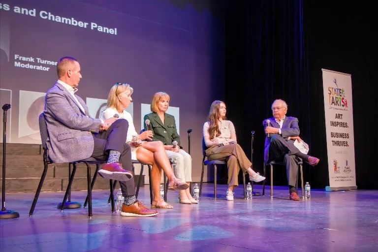 Five people sitting on stage participating in a panel discussion, with a screen behind them displaying text and a banner to the right that reads "State Fair" and promotes art, inspiration, business, and thriving.