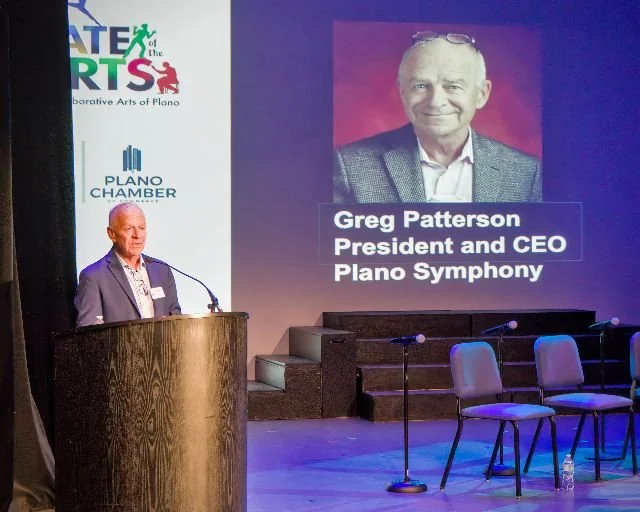 A man standing at a podium speaking at an event, with a large screen behind him displaying a photo and text introducing Greg Patterson, President and CEO of Plano Symphony, at an arts event at the Plano Chamber.