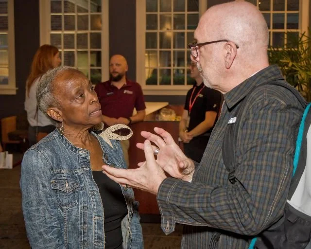 An elderly woman with short gray hair and a denim jacket engaging in a conversation with a bald man wearing glasses and a plaid shirt in a social setting.