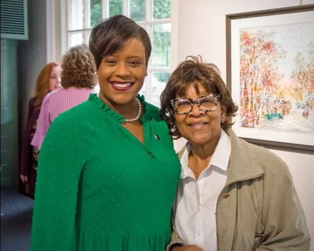 Two women smiling indoors near a window and artwork, with other people in the background.