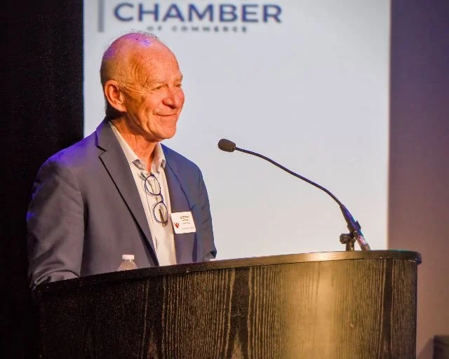 An older man in a suit stands at a podium with a microphone, smiling during a speech or presentation at a formal event.