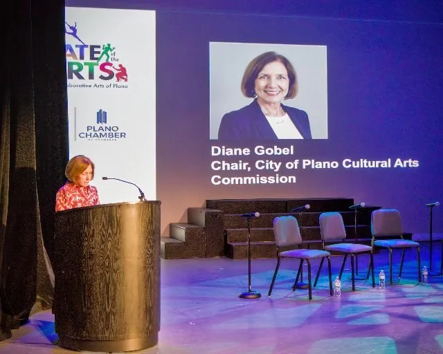 A woman standing at a wooden podium giving a speech at an event. Behind her, a large screen displays her photo and title: "Diane Gobel, Chair, City of Plano Cultural Arts Commission." The stage has four empty chairs and a water bottle on the floor.