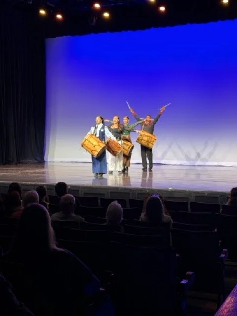 Four performers on stage holding traditional drums and sticks, with audience members watching in a theater.