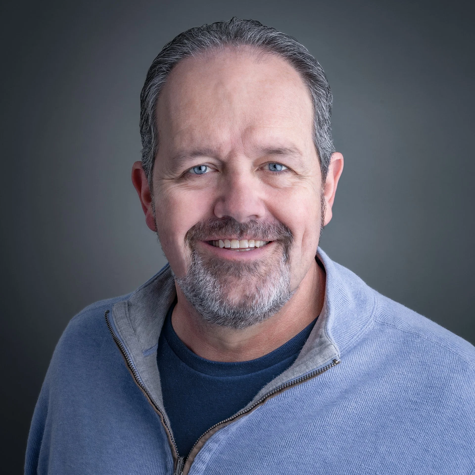 A person smiling outdoors, wearing a red jacket and pearl necklace with a blurred background of a brick building.