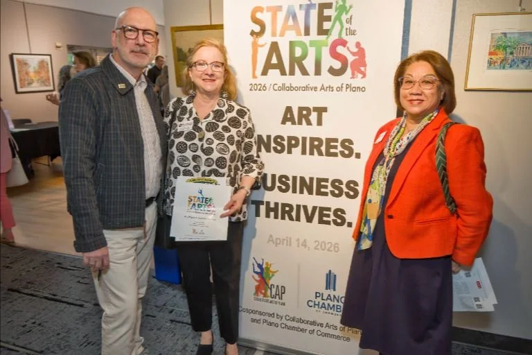 Three people standing indoors in front of a banner at the State of the Arts event, April 14, 2026. The man on the left is wearing glasses, a dark checkered blazer, and light-colored pants. The woman in the middle has blonde hair, glasses, and is dres