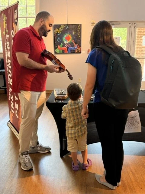 A man playing a violin for a woman and a young boy at an indoor event, with colorful artwork on the wall behind them.