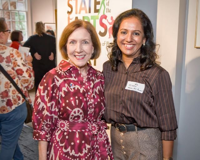 Two women smiling at a professional event, standing in front of a sign that reads "State of the Arts." One woman has short red hair and is wearing a pink and maroon patterned dress; the other has long dark hair and is wearing a brown striped blouse a