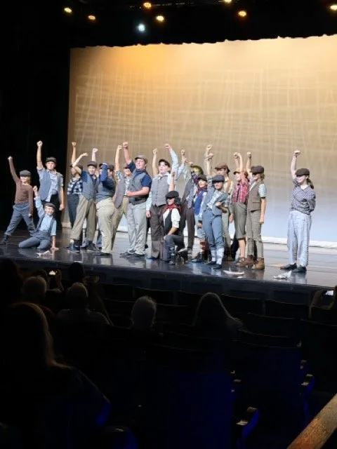 Children dressed in vintage clothing on stage with some raising their fists, performing in front of an audience.