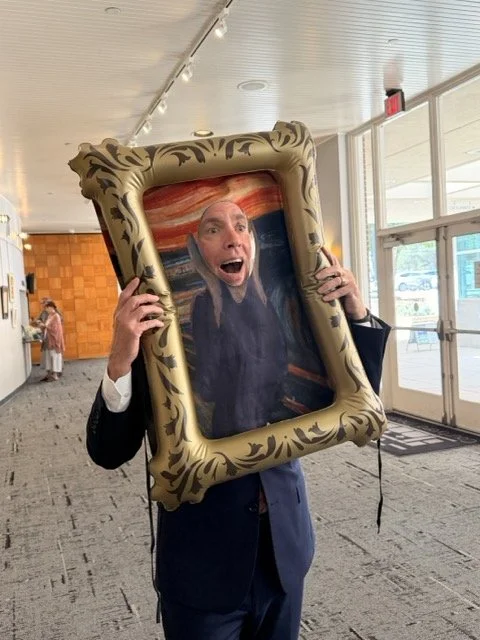 Person holding a large picture frame with a painted portrait of a man with surprised expression on his face.