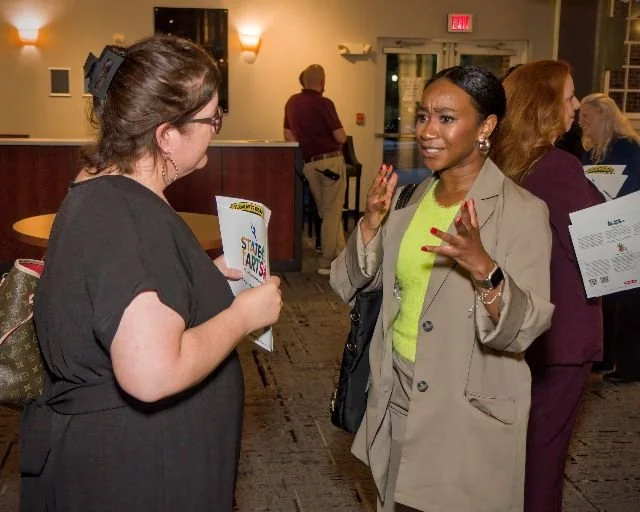 Two women engage in conversation at a social event inside a building, one holding a flyer.