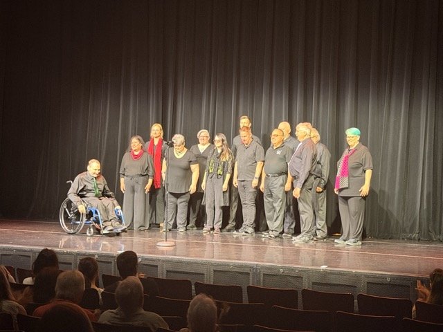 Group of people on stage in front of a black curtain, with some in wheelchairs, posing for a photo, audience in the foreground.