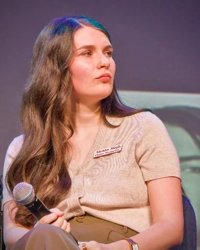A young woman with long, wavy brown hair, wearing a beige short-sleeve cardigan, holding a microphone, during a panel or conference.