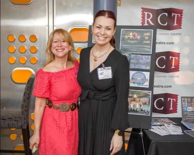 Two women standing indoors, smiling at the camera. The woman on the left wears a red dress with a wide belt, and the woman on the right wears a black dress with a necklace and a name badge. Behind them is a display board with posters and a banner for