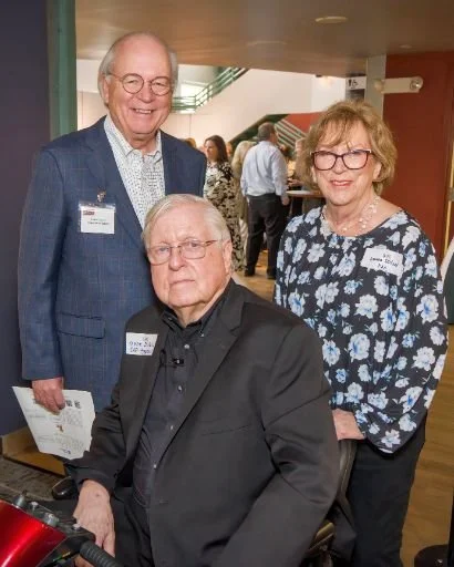 Three older adults at a social event, two men and one woman, standing and sitting together indoors, smiling, wearing name tags.