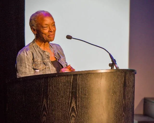 An elderly woman standing behind a wooden podium with a microphone, appearing to be speaking or giving a speech at an indoor event.