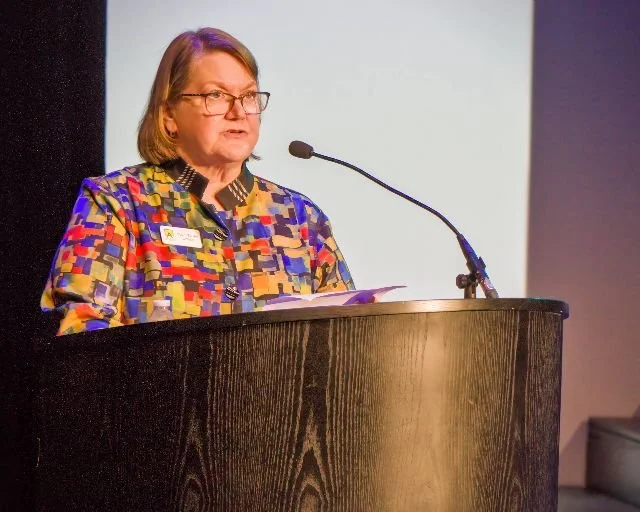 A woman with glasses, wearing a colorful, patterned jacket, speaking at a podium with a microphone during a presentation or speech.