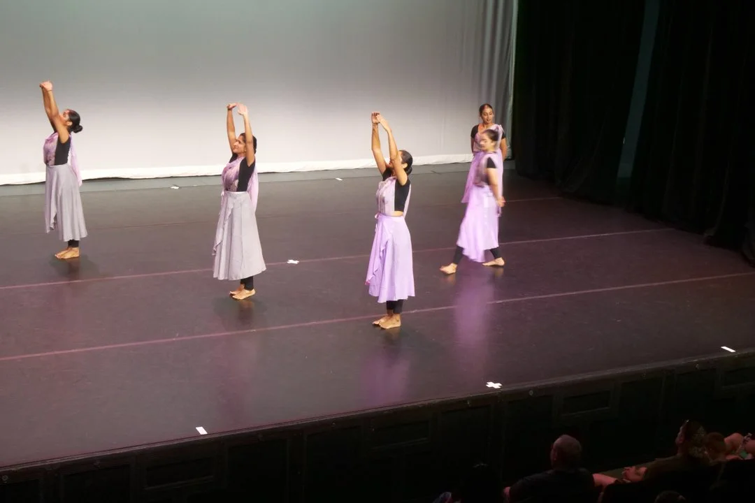 Five women performing a dance routine on stage, wearing matching purple and black traditional-style dresses with black tops and purple skirts, some with long skirts and some with shorter skirts, in front of a plain white backdrop, with audience membe