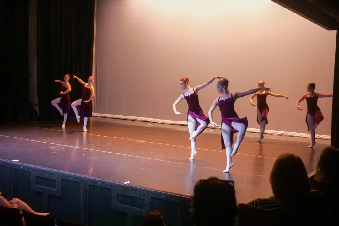 Ballet dancers perform on stage in purple costumes with a plain backdrop.