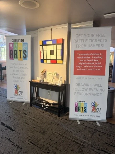 Indoor exhibition area with two banners and a small table. The left banner promotes Plano's annual arts festival, while the right banner advertises a raffle for free tickets from Ushers, with details about prizes and conditions. A colorful geometric 