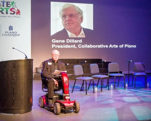 An elderly man with glasses wearing a suit and blazer, sitting on a red mobility scooter, reading a document on a stage with blank chairs, a podium, and a large screen behind him displaying a photo of Gene Dillard, President of Collaborative Arts of 
