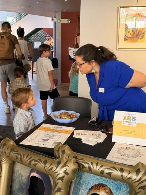 A woman in a blue uniform leaning over a table to talk to a young boy with red hair and a patterned shirt at a busy indoor event. The table has papers, a bowl of food, and a sign that reads '105 Show 2020'. People are walking in the background near a