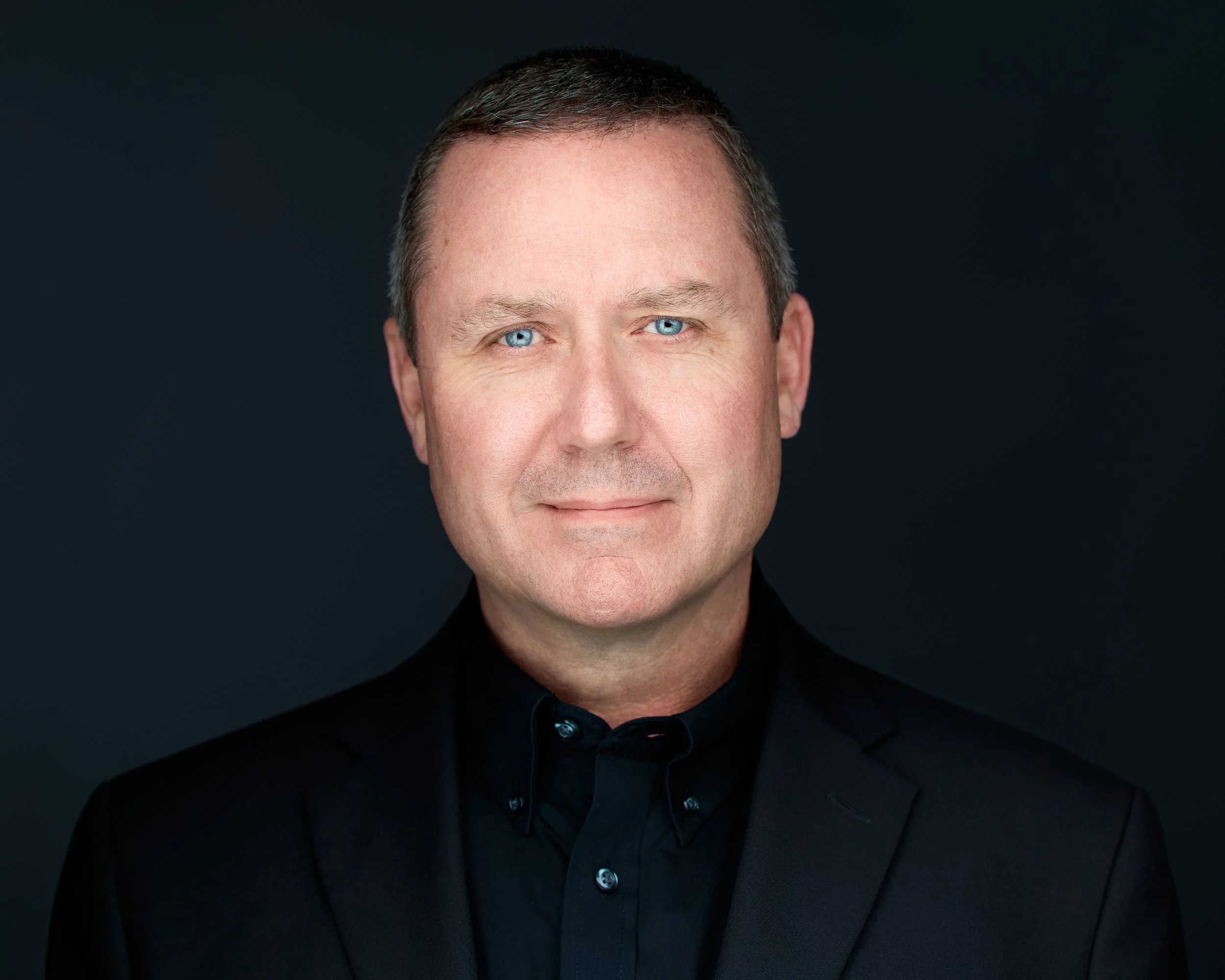 A smiling woman with long wavy hair wearing a black top against a neutral background.
